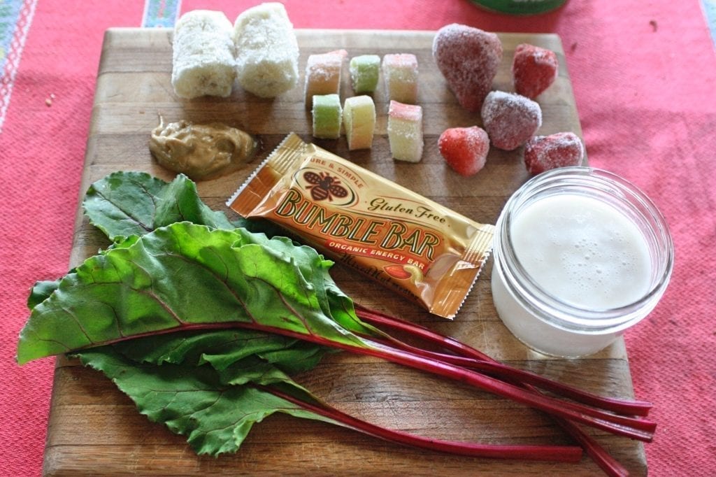 A Mixed Nut Medley BumbleBar is the featured smoothie ingredient on this wooden cutting board with banana, rhubarb, strawberry, almond milk, cashew butter, and beet greens.