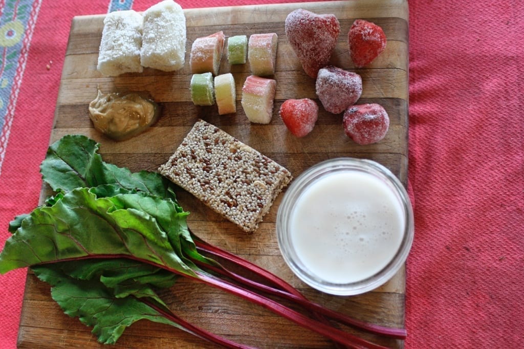 A Mixed Nut Medley BumbleBar is the featured smoothie ingredient on this wooden cutting board with banana, rhubarb, strawberry, almond milk, cashew butter, and beet greens.