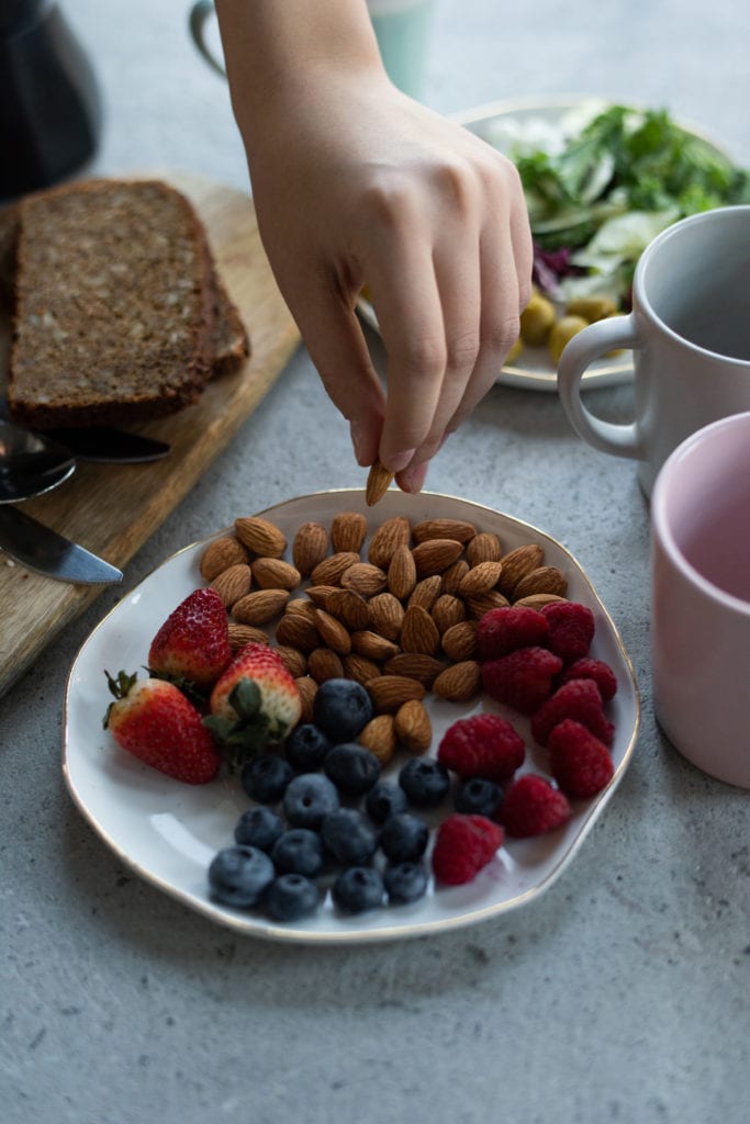 Person holding organic almond above a plate of almonds, strawberries, blueberries, and raspberries